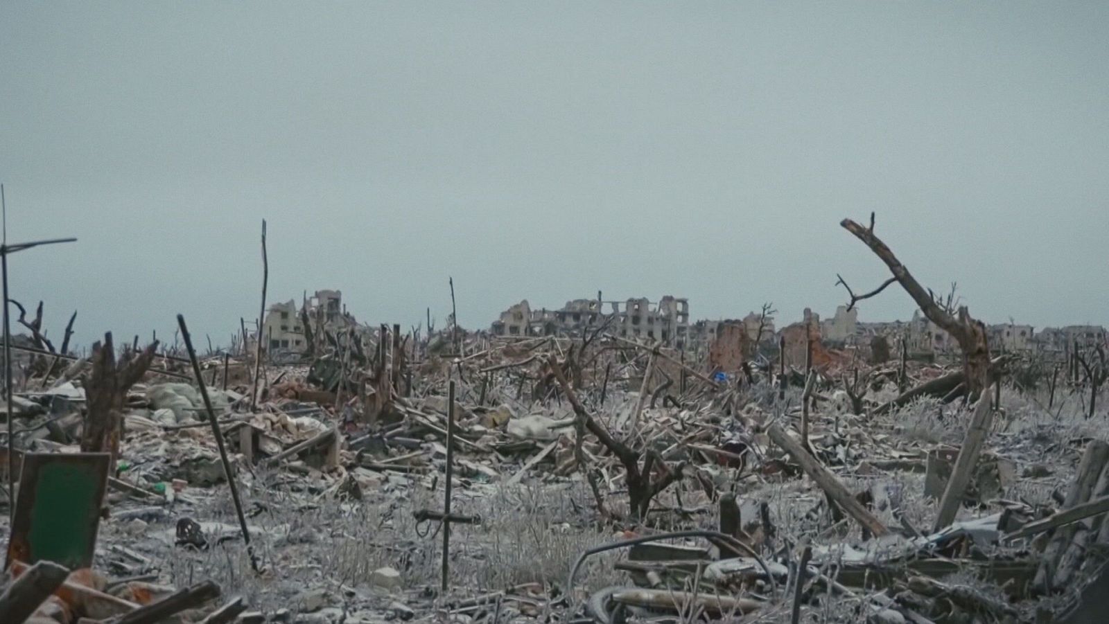 Una escena de destrucción muestra edificios en ruinas y escombros, con restos dispersos por el suelo y vegetación dañada, dominada por tonos grises que evocan una atmósfera de desolación.