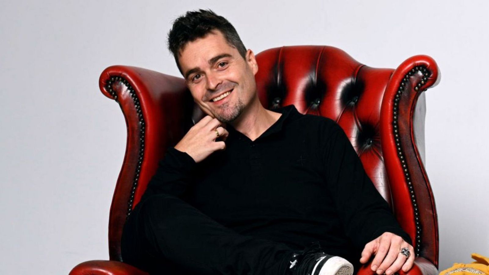 Un hombre con barba y canas, sonríe a la cámara sentado en un sillón rojo. Viste ropa oscura y lleva anillos en las manos.