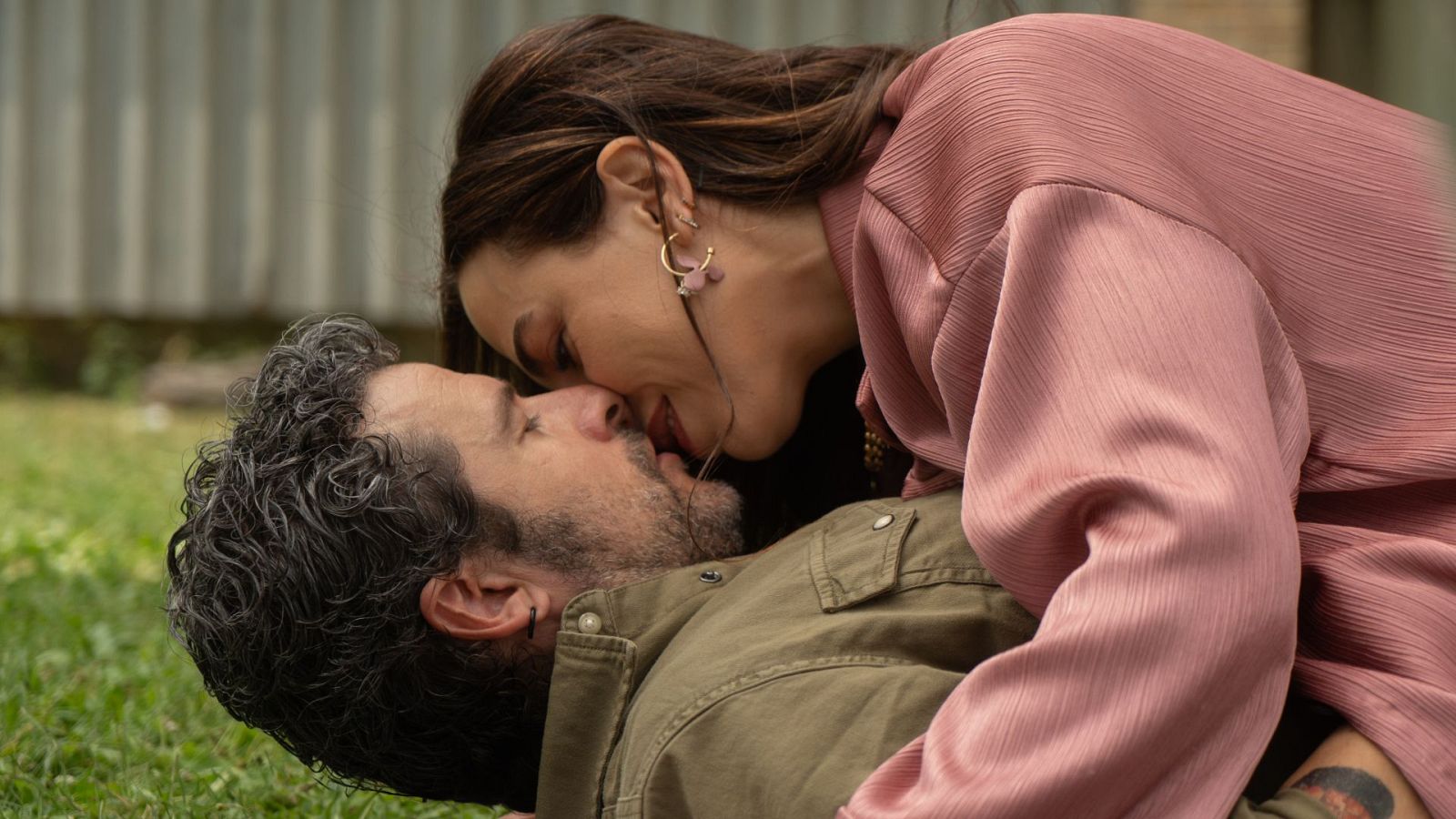 Una pareja, Ricardo y Sonia, comparte un beso apasionado en un entorno al aire libre. Él viste una camisa verde oliva y está tumbado, mientras ella, con una blusa rosa, se encuentra encima.