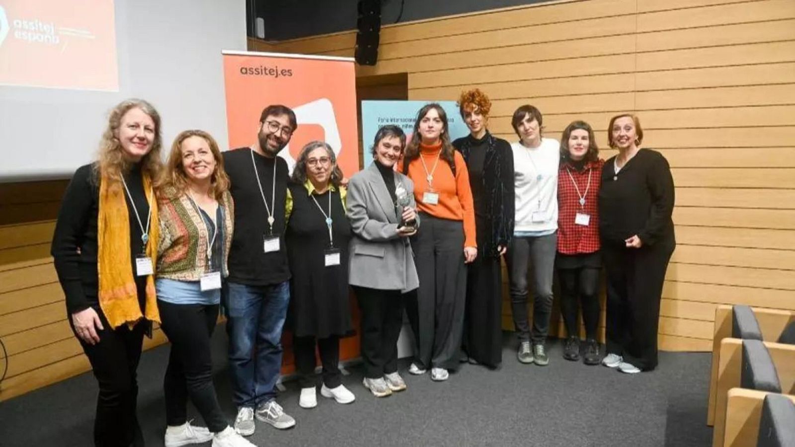 Nueve personas, entre hombres y mujeres, posan en un espacio interior. Visten ropa variada y llevan identificaciones. Se observan carteles al fondo y sillas a la derecha.