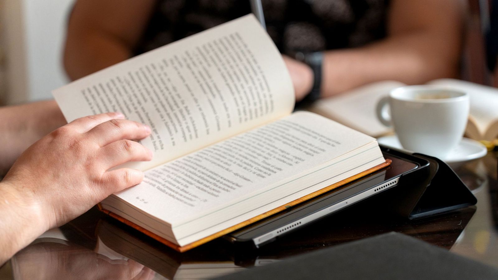 Una persona lee un libro abierto, apoyado sobre una tablet. Se observa una taza de café y otra persona en el fondo, sobre una mesa oscura.