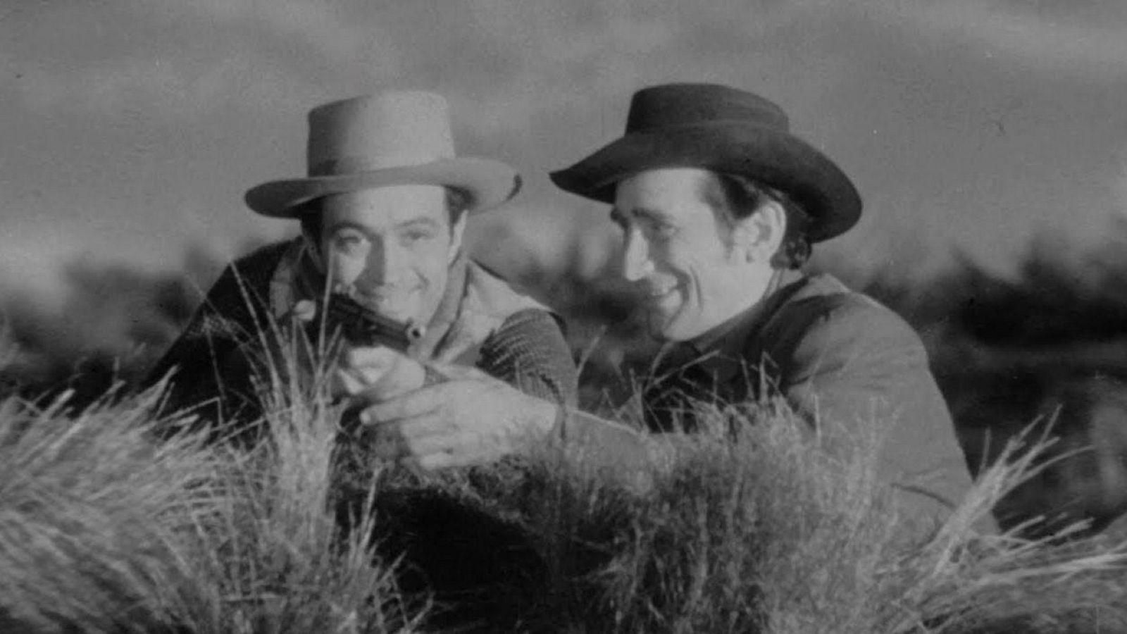 En una escena en blanco y negro, dos hombres agachados en un campo, uno con sombrero claro y chaleco empuñando un revólver, y el otro con sombrero oscuro mirando en la misma dirección, posiblemente en un ambiente del oeste.