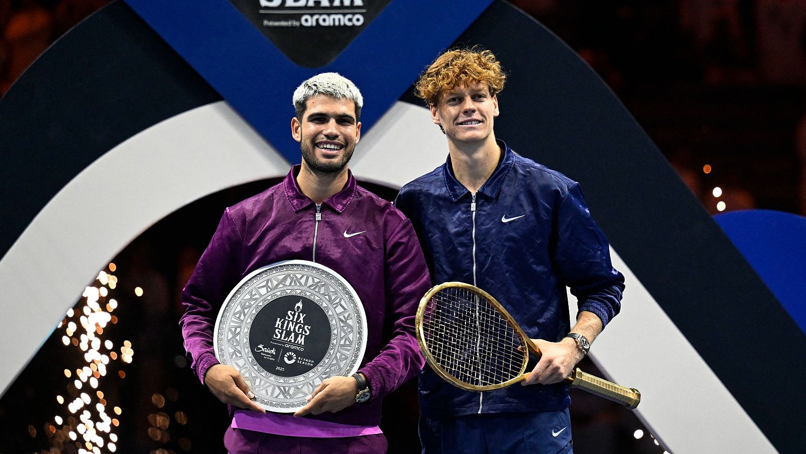 Dos tenistas, uno con el pelo rubio y otro pelirrojo, posan juntos. Uno de ellos sostiene un plato plateado y el otro una raqueta dorada, ambos con chaquetas deportivas.