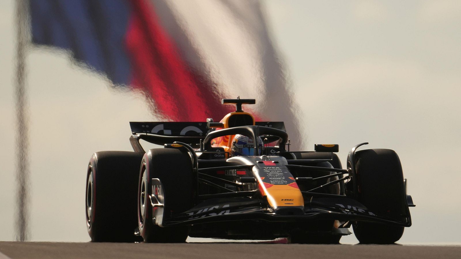 Un coche de Fórmula 1 negro con detalles en naranja y azul, con el piloto dentro, y una bandera ondeando al fondo, bajo un cielo despejado.