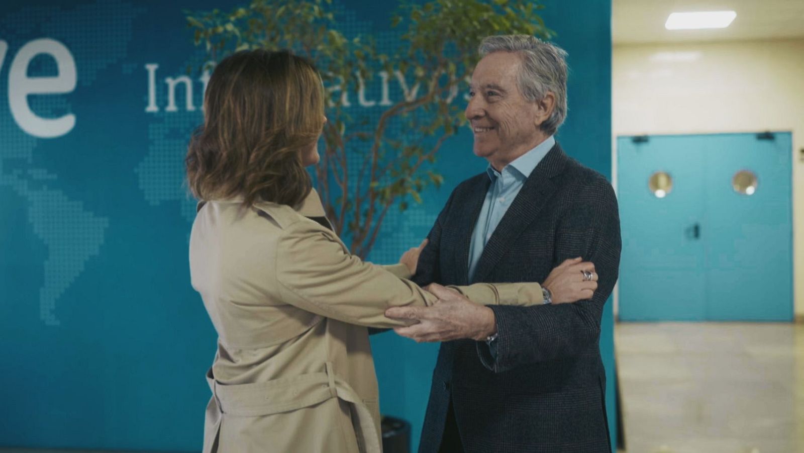 En un plató de televisión, Iñaki Gabilondo saluda a una mujer frente a un panel con el logotipo de "RTVE Informativos" y un mapa del mundo. Se observa una planta decorativa y un pasillo con una puerta.