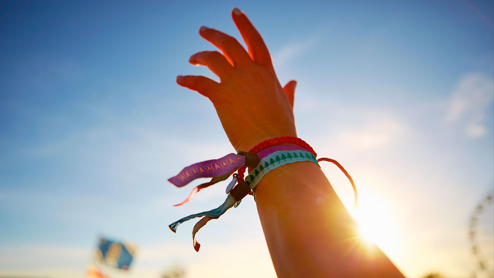 Una mano con pulseras de colores se alza hacia el cielo soleado, con una bandera y una noria al fondo, evocando un ambiente de festival.