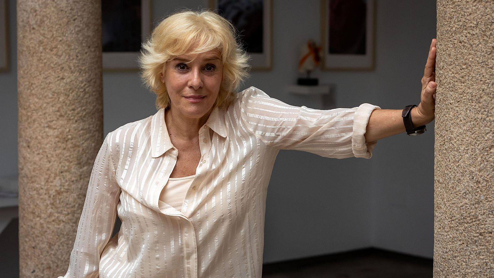 Anabel Alonso, con camisa a rayas, se apoya en una columna de piedra. Se observa una escultura y cuadros en el fondo, en un ambiente que parece ser una galería.