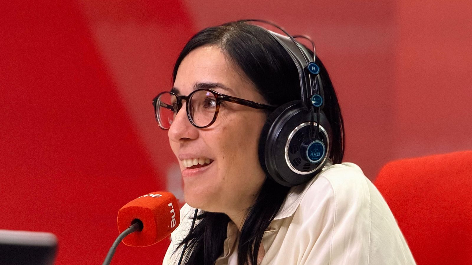 Una mujer con gafas y auriculares AKG sonríe frente a un micrófono rojo, posiblemente en un estudio de grabación. El fondo es de color rojo.