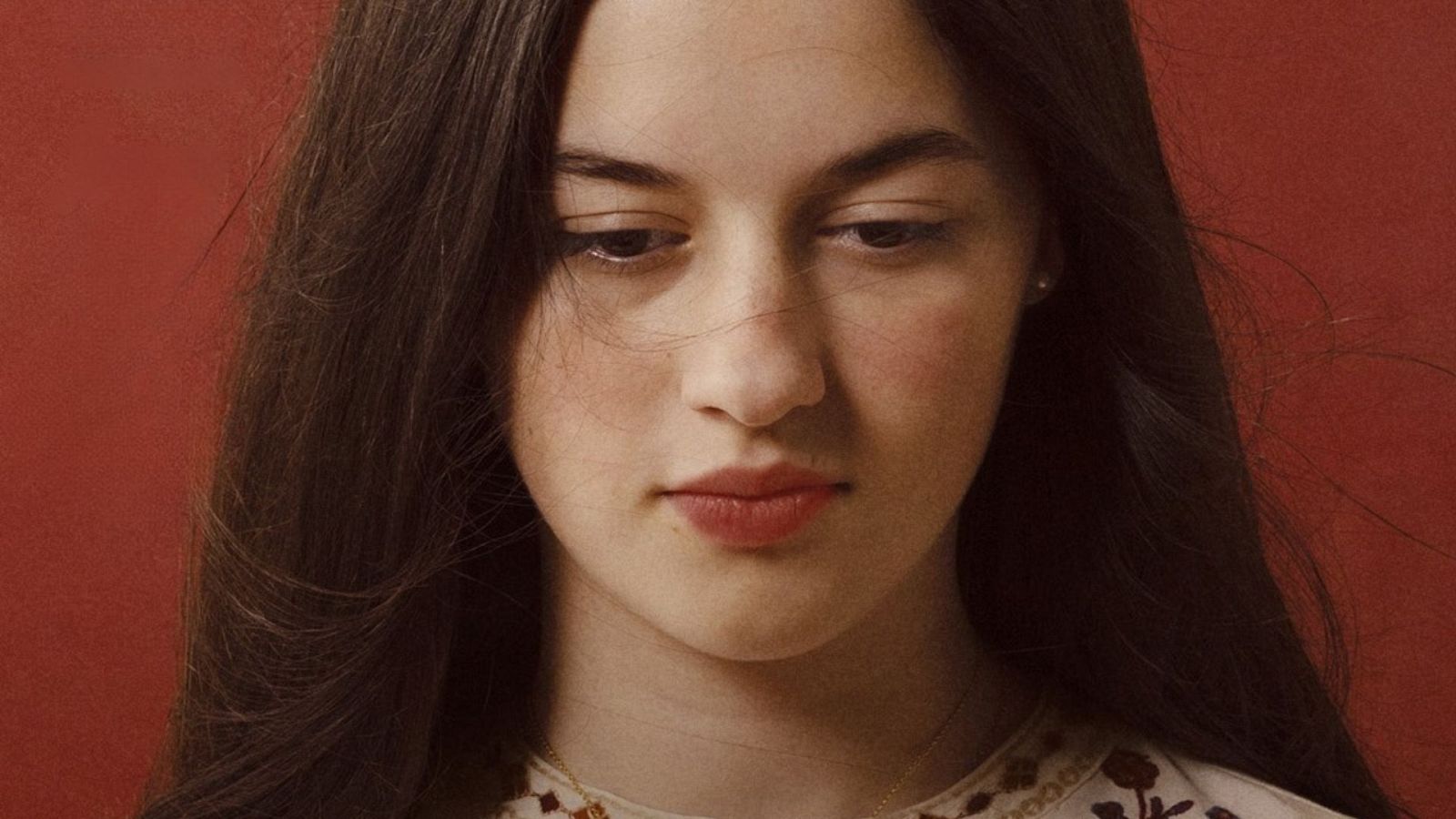 Blanca Soroa, con cabello castaño oscuro y liso, luce un collar dorado y una blusa blanca con bordados, mientras mira hacia abajo. El fondo es de color rojo oscuro.