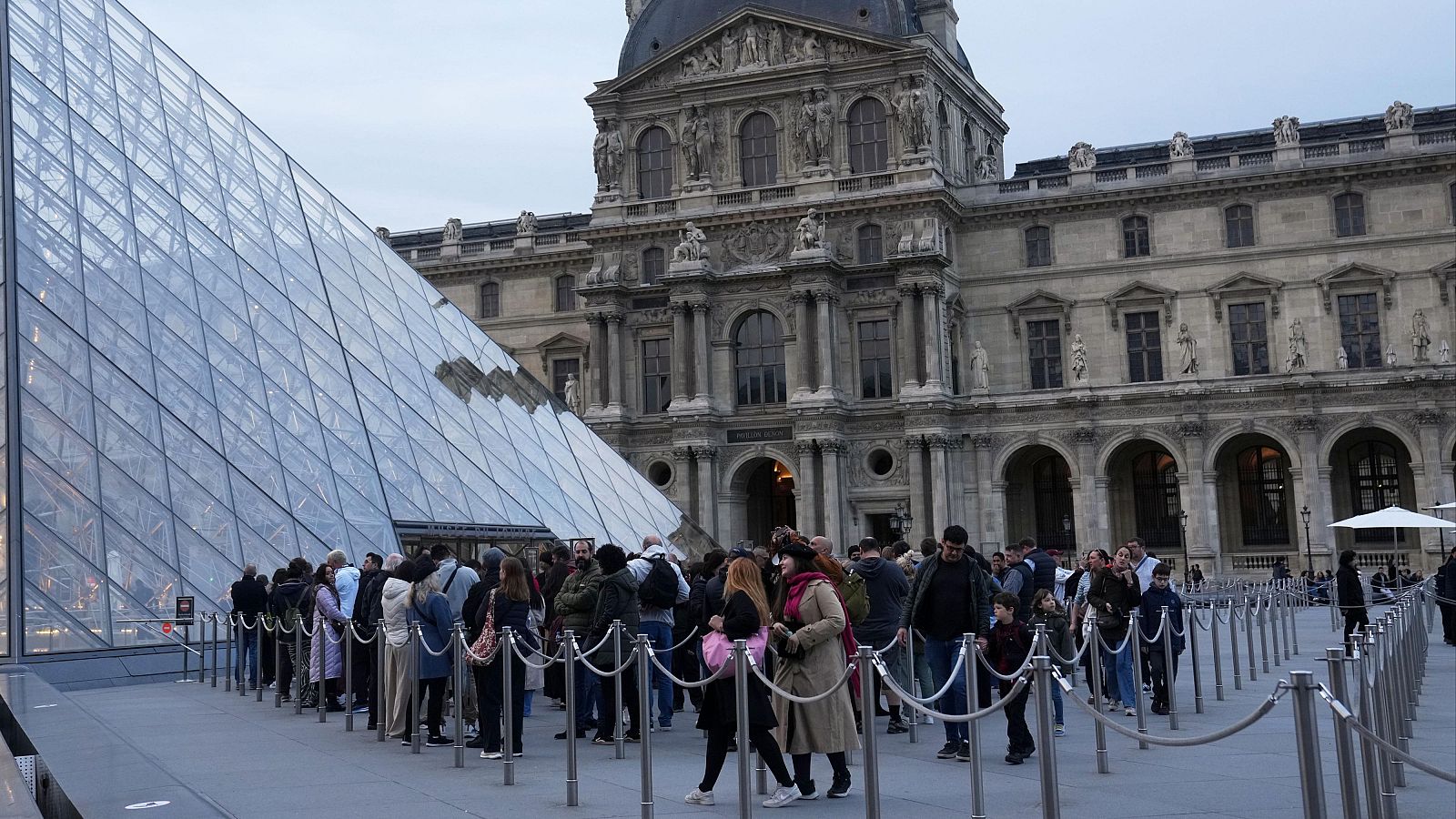 En primer plano, la pirámide de cristal del Louvre se alza frente al edificio principal del museo, con una fila de personas esperando para entrar, delimitada por barreras.