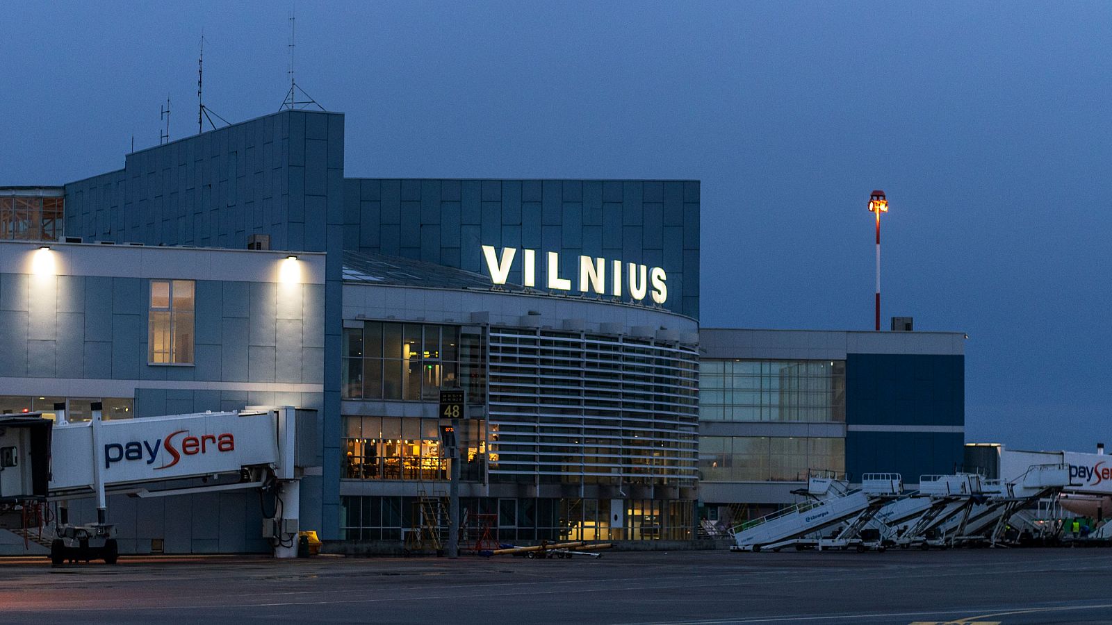 En la imagen se aprecia el aeropuerto, con su edificio principal de diseño moderno y la palabra "VILNIUS" destacada. Se observan pasarelas de embarque, una de ellas con una marca visible, en un ambiente con iluminación tenue y cielo oscuro.