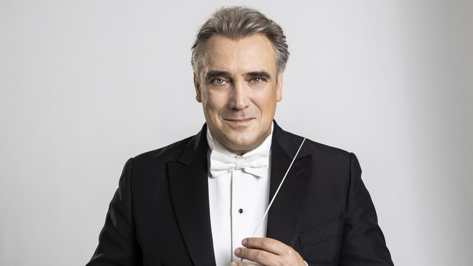 Jaime Martín, con cabello canoso y peinado hacia atrás, posa con un esmoquin negro, camisa blanca y pajarita. Sostiene una batuta blanca, mostrando una expresión serena.