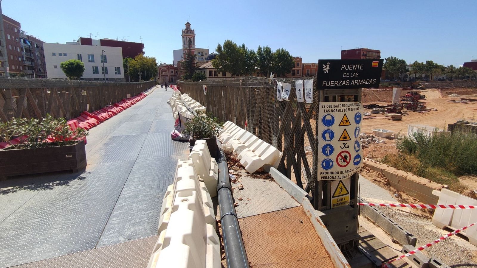 Un puente provisional de metal, con señalización de seguridad y barreras, atraviesa un barranco. Se observa una señal que indica el nombre del puente y símbolos de seguridad. Al fondo, se distinguen edificios y una torre.