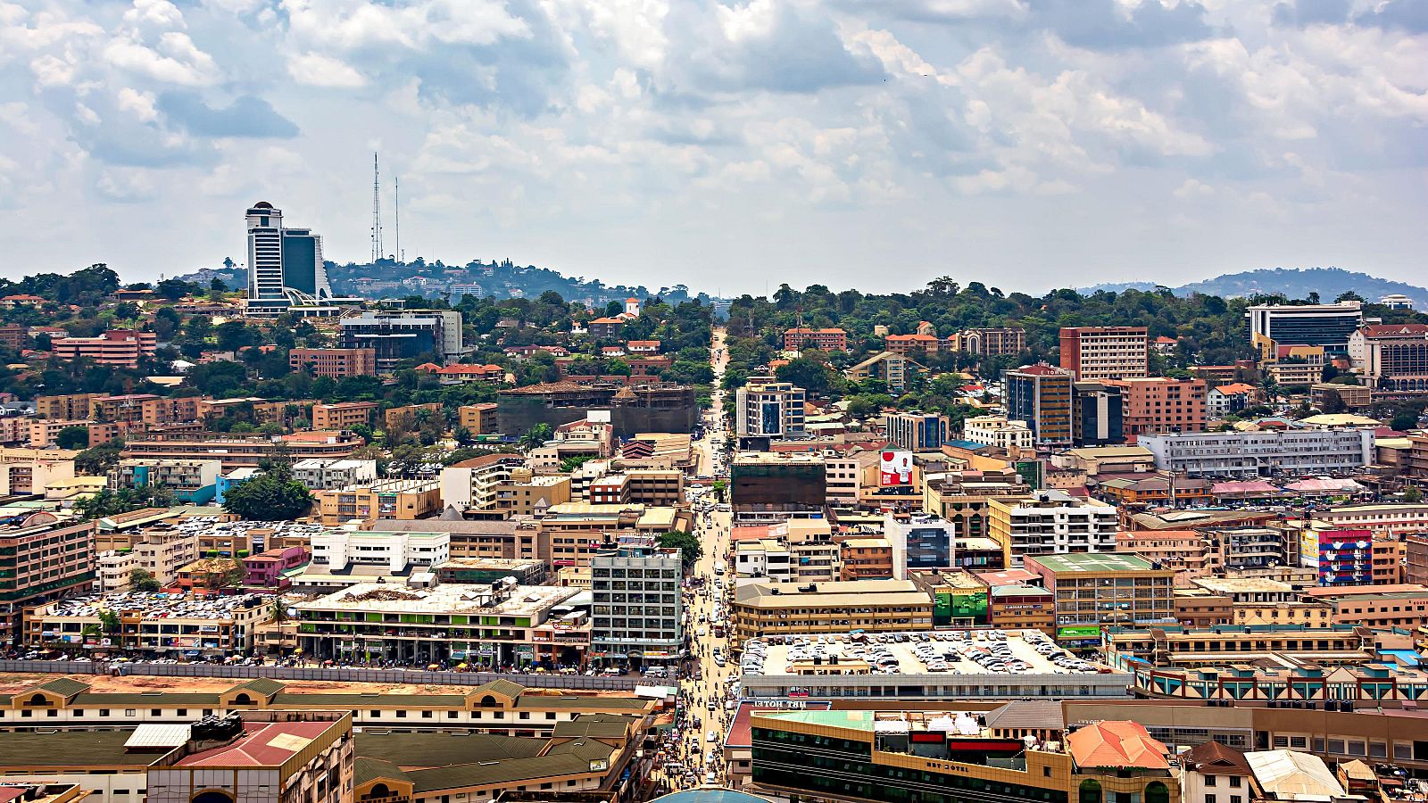 Una panorámica de Kampala, capital de Uganda, revela una ciudad bulliciosa con edificios variados y una calle principal con tráfico, contrastando con colinas verdes y un cielo parcialmente nublado.