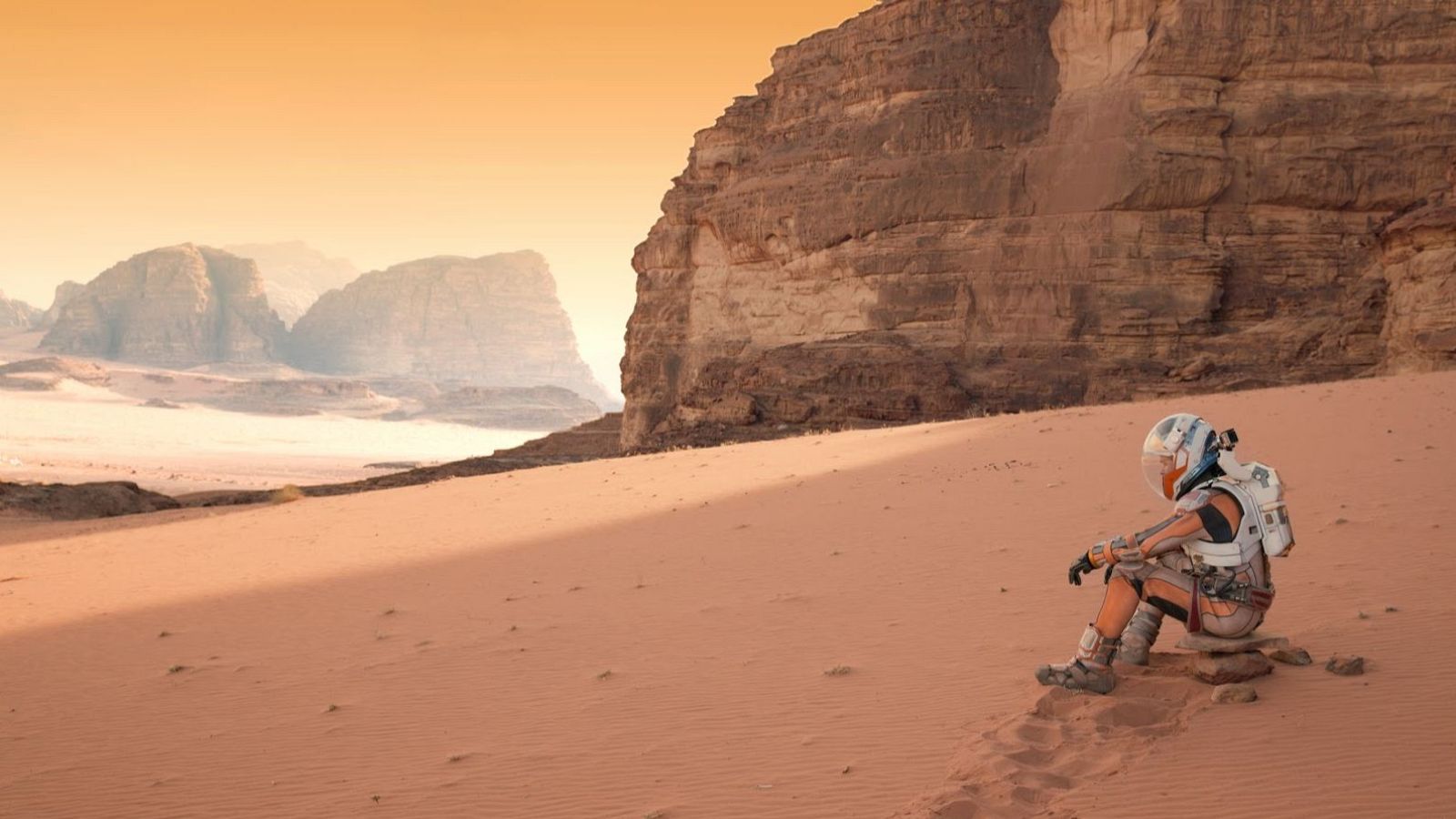 Un astronauta solitario, vestido con un traje espacial blanco y naranja, descansa sobre rocas en un entorno desértico de color rojizo, con formaciones rocosas y un cielo anaranjado.