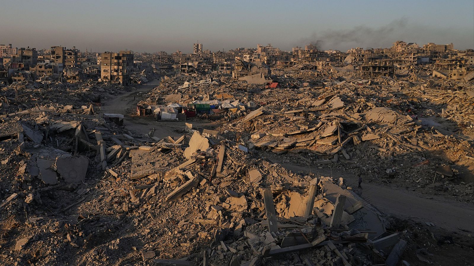 Un paisaje urbano desolado en una ciudad, con edificios en ruinas y escombros por todas partes. Se observa humo en la distancia y posibles refugios improvisados, evidenciando la magnitud de la devastación.