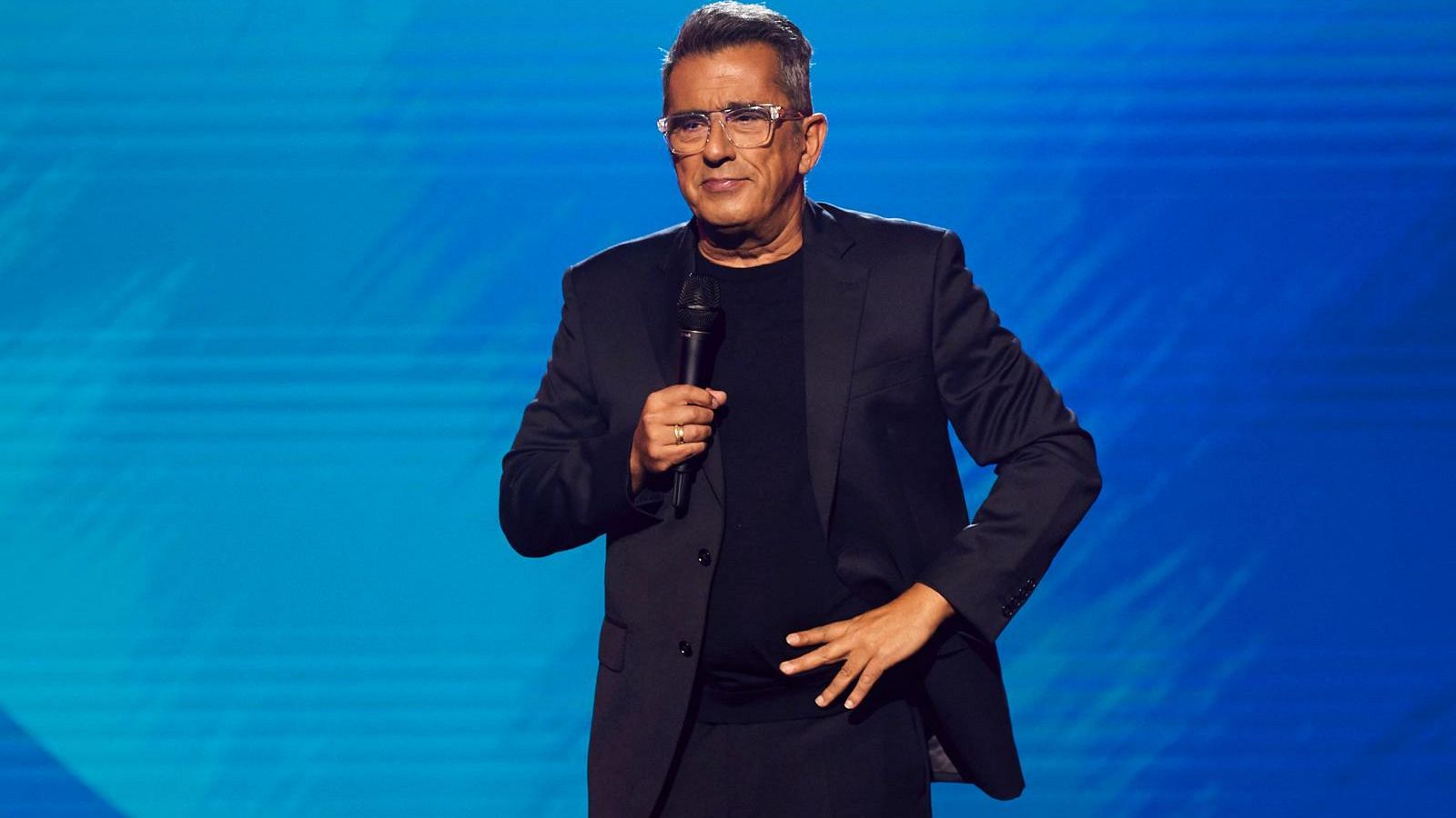 En un escenario con fondo azul, Andreu Buenafuente, vestido de negro, sostiene un micrófono mientras posa con una mano en la cadera.