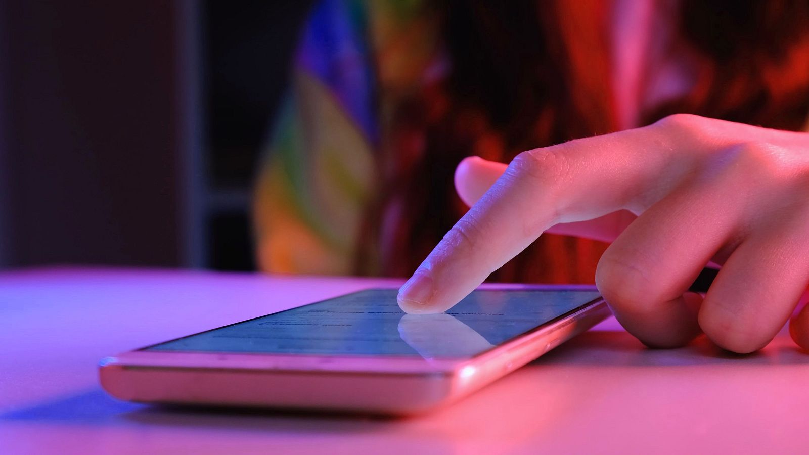 Una mano toca la pantalla de un teléfono móvil, que se encuentra sobre una superficie iluminada con luces rosas y azules. Al fondo, se distingue una bandera del arcoíris, creando un ambiente vibrante.