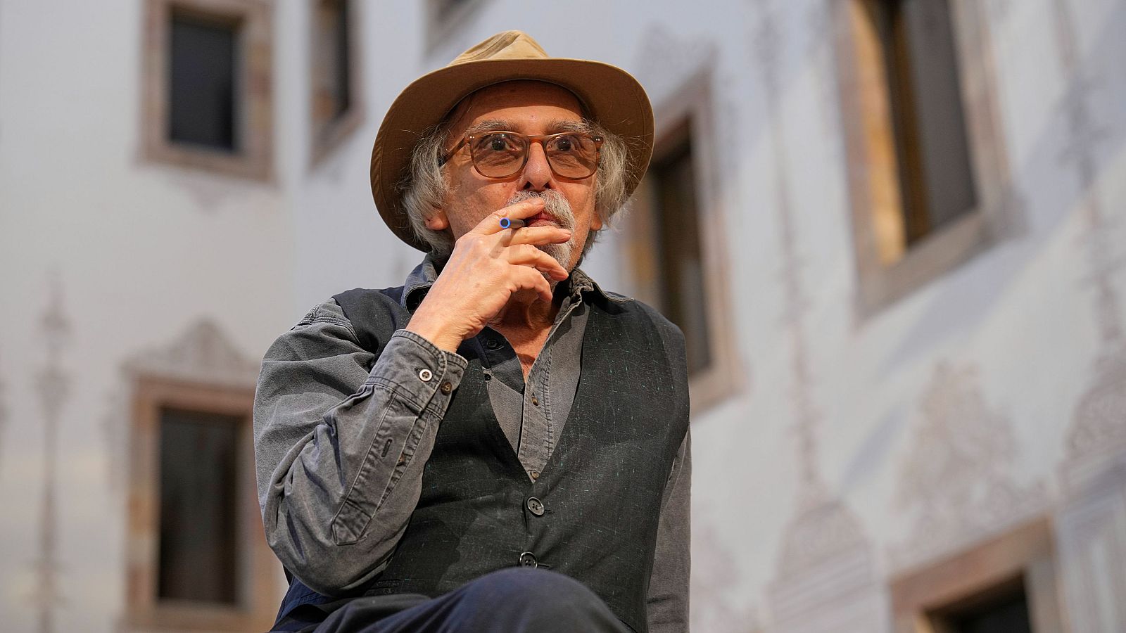 Art Spiegelman, con sombrero y gafas, fuma con la mano cerca de la boca, vistiendo una camisa gris y un chaleco oscuro, con un edificio de fondo.