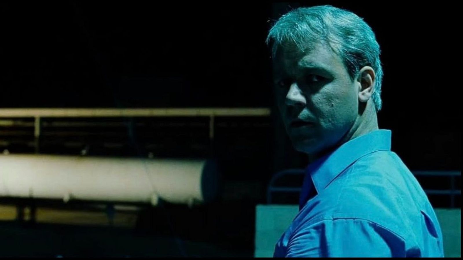 En primer plano, un hombre de mediana edad con cabello canoso y camisa azul muestra una expresión seria. El fondo oscuro presenta una estructura metálica horizontal, con una iluminación tenue y un ligero tono azulado.