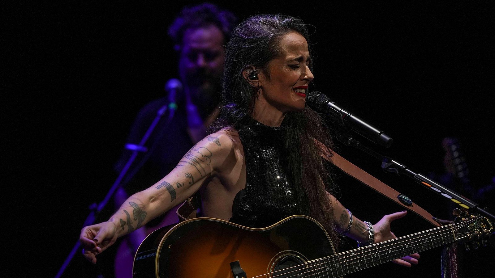 Vega, con cabello oscuro y canas, interpreta en un escenario con un vestido negro brillante y tatuajes, acompañada de una guitarra y un micrófono, mientras otros músicos la acompañan.