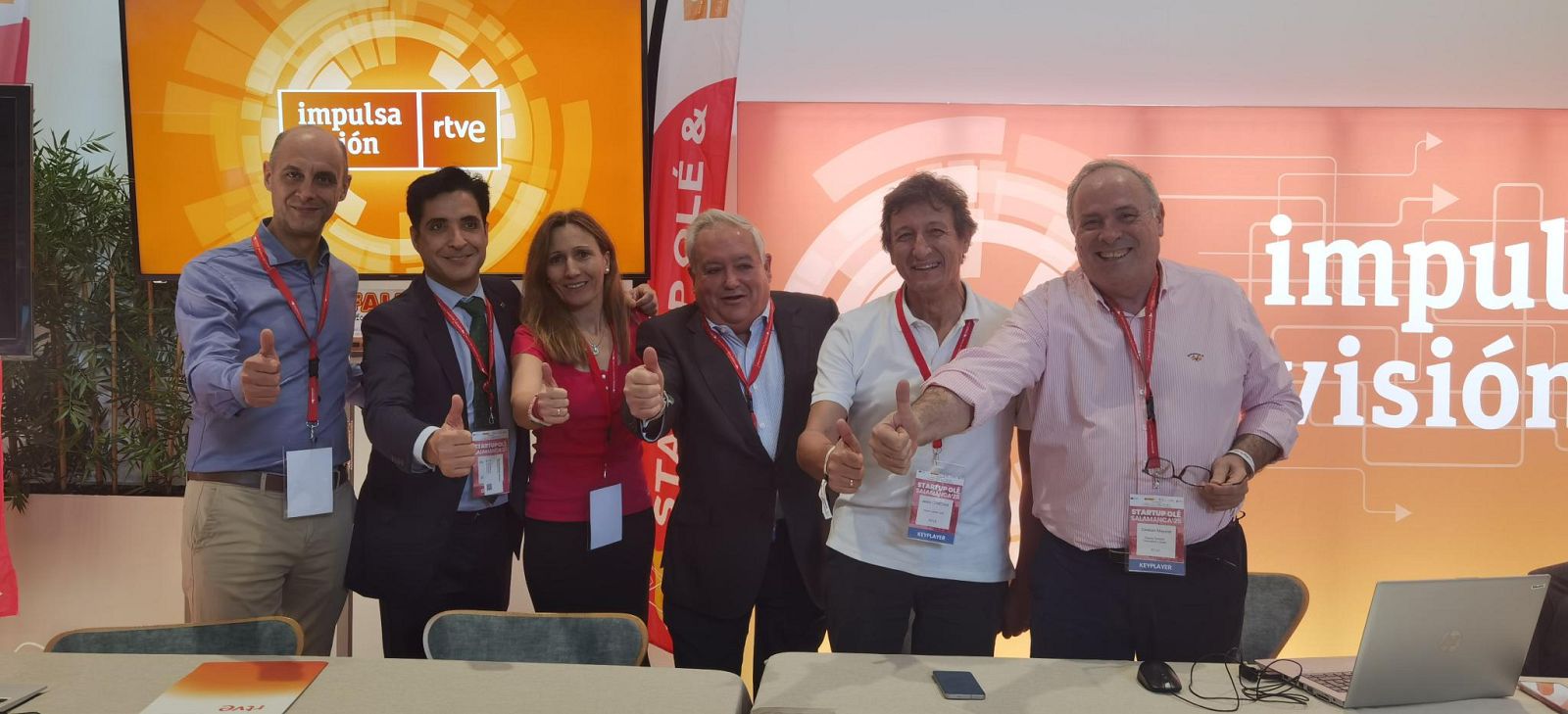 Seis personas sonríen y hacen el gesto de aprobación frente a una pantalla con logos y un cartel, rodeados de ordenadores portátiles y teléfonos.