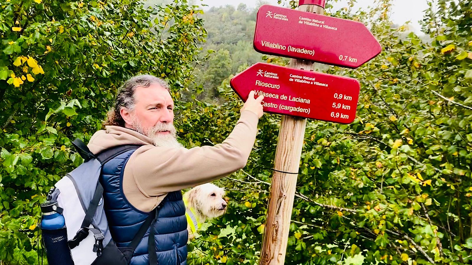 Juan señala un cartel de dirección en el Camino Natural Vía Verde de Laciana junto a su perro Migas, rodeados de vegetación.