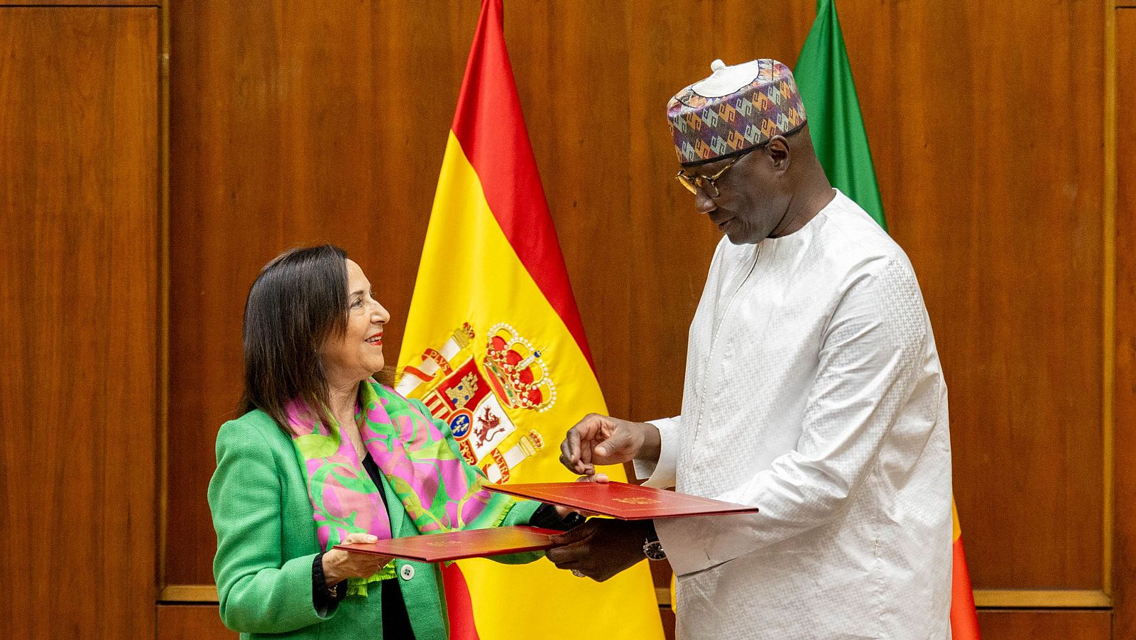 Encuentro entre Margarita Robles y Birame Diop, donde intercambian documentos. La ministra española viste una chaqueta verde y una bufanda floral, mientras que su homólogo senegalés lleva un traje blanco y un gorro.