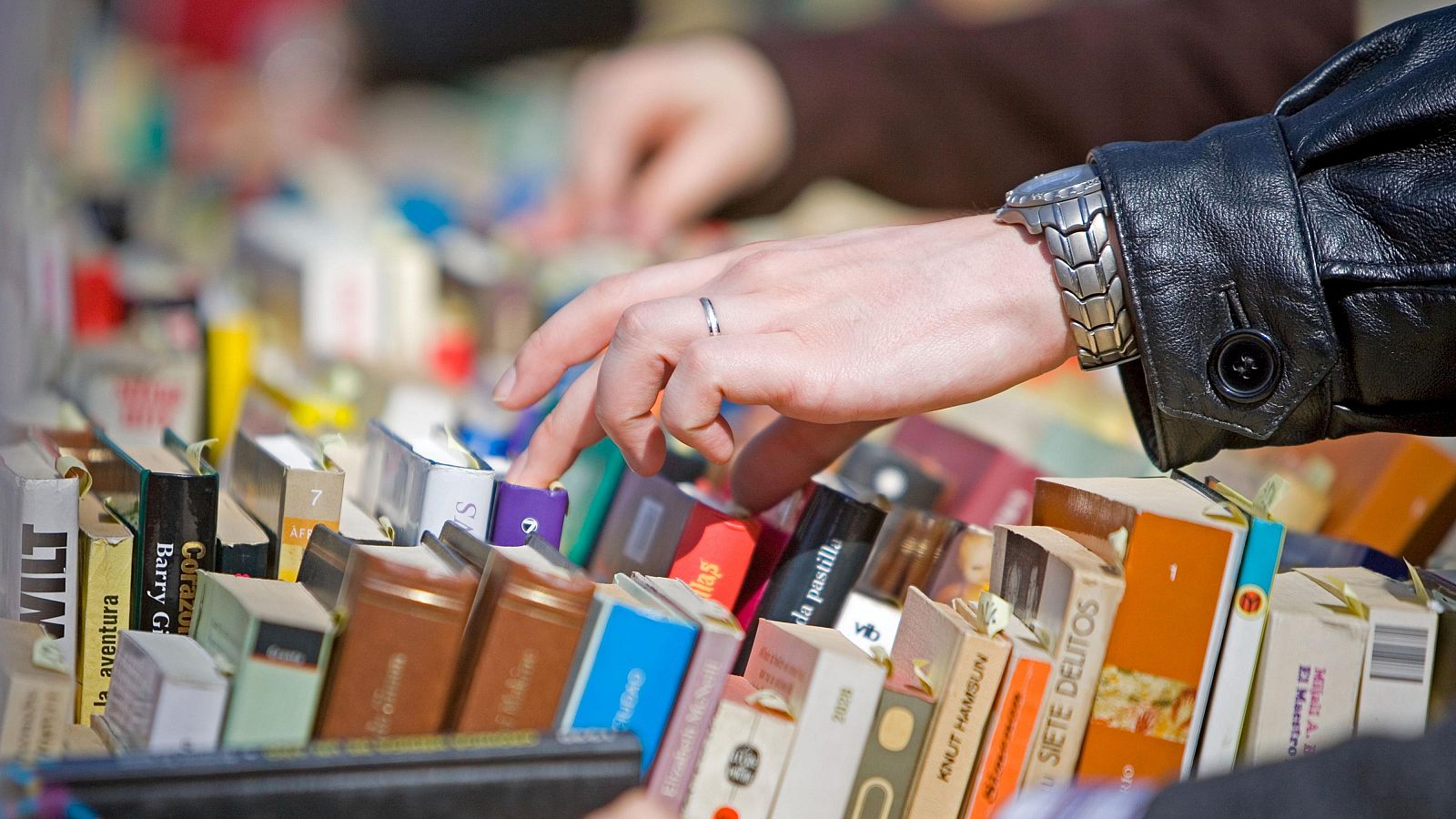 Una mano con un anillo plateado explora una pila de libros de bolsillo, posiblemente buscando un título específico. Se aprecia un reloj de pulsera con correa metálica en la muñeca y una chaqueta de cuero negra.