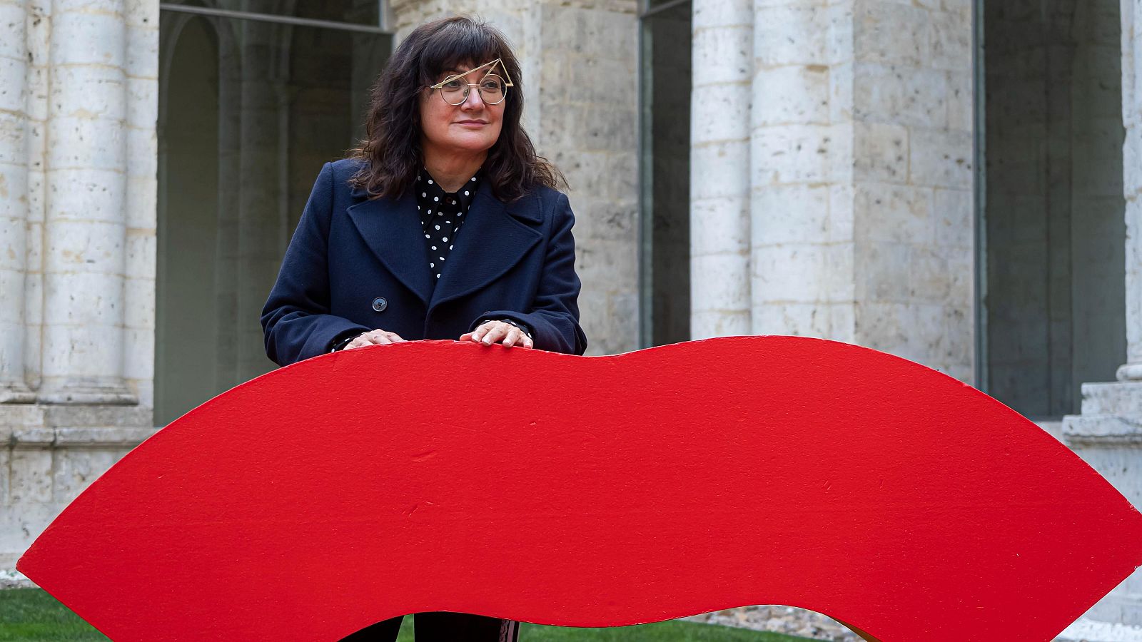 Isabel Coixet, con gafas y abrigo azul marino, sostiene una gran escultura roja con forma de labios, frente a una arquitectura de piedra clara.