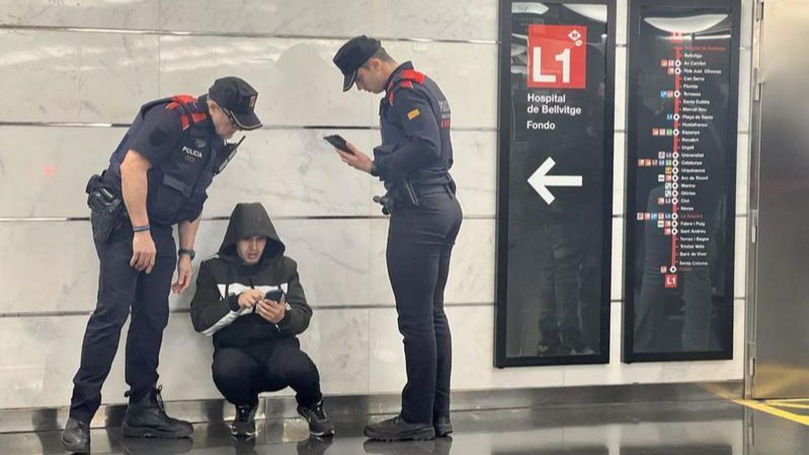En una estació de metro, dos policies uniformats interactuen amb un jove que està agachado mirant el seu telèfon. Un cartell amb el mapa de la línia L1 del metro de Barcelona és visible al fons.