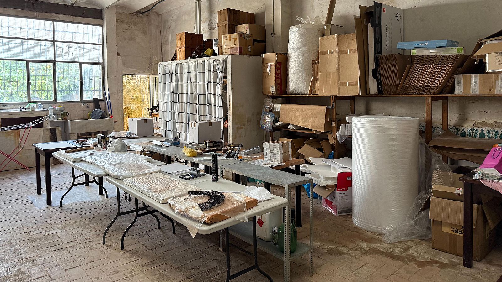 Interior de un taller artesanal con mesas de trabajo repletas de materiales y objetos, posiblemente relacionados con la fabricación de abanicos. Se aprecia una estantería con cajas y una ventana que ilumina el espacio.