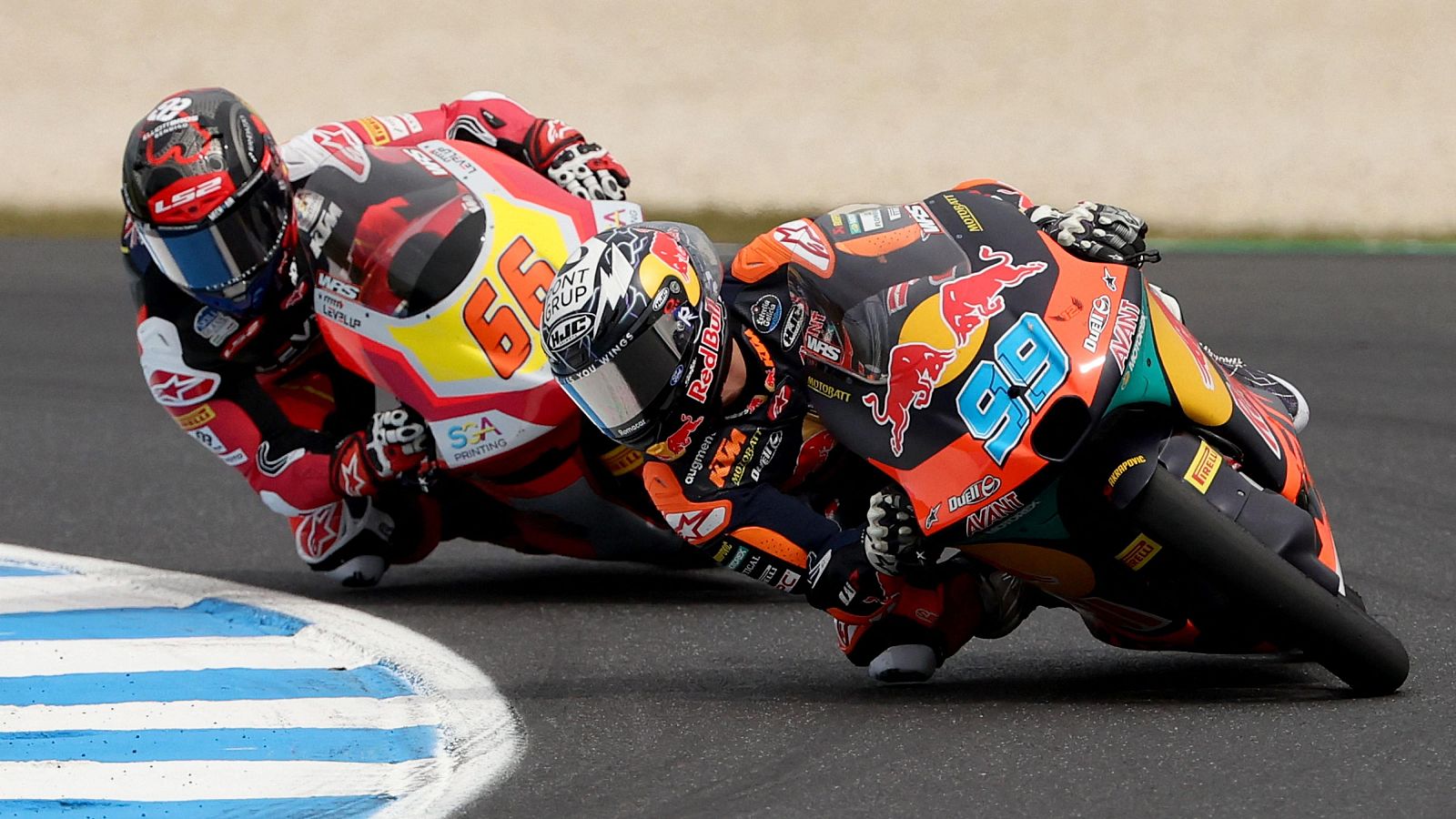 Dos pilotos de Moto3 compiten en una curva, uno con el número 99 y equipación naranja, y otro con el número 66 y equipación roja, ambos inclinados hacia la izquierda en plena carrera.