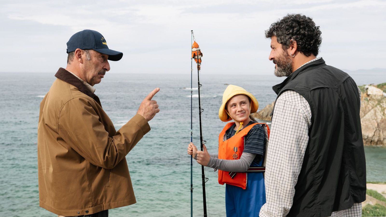 Un fotograma de una película muestra a tres personas en la costa. Un hombre con gorra y chaqueta señala, un niño con sombrero y chaleco sostiene una caña de pescar, y otro hombre con barba observa.