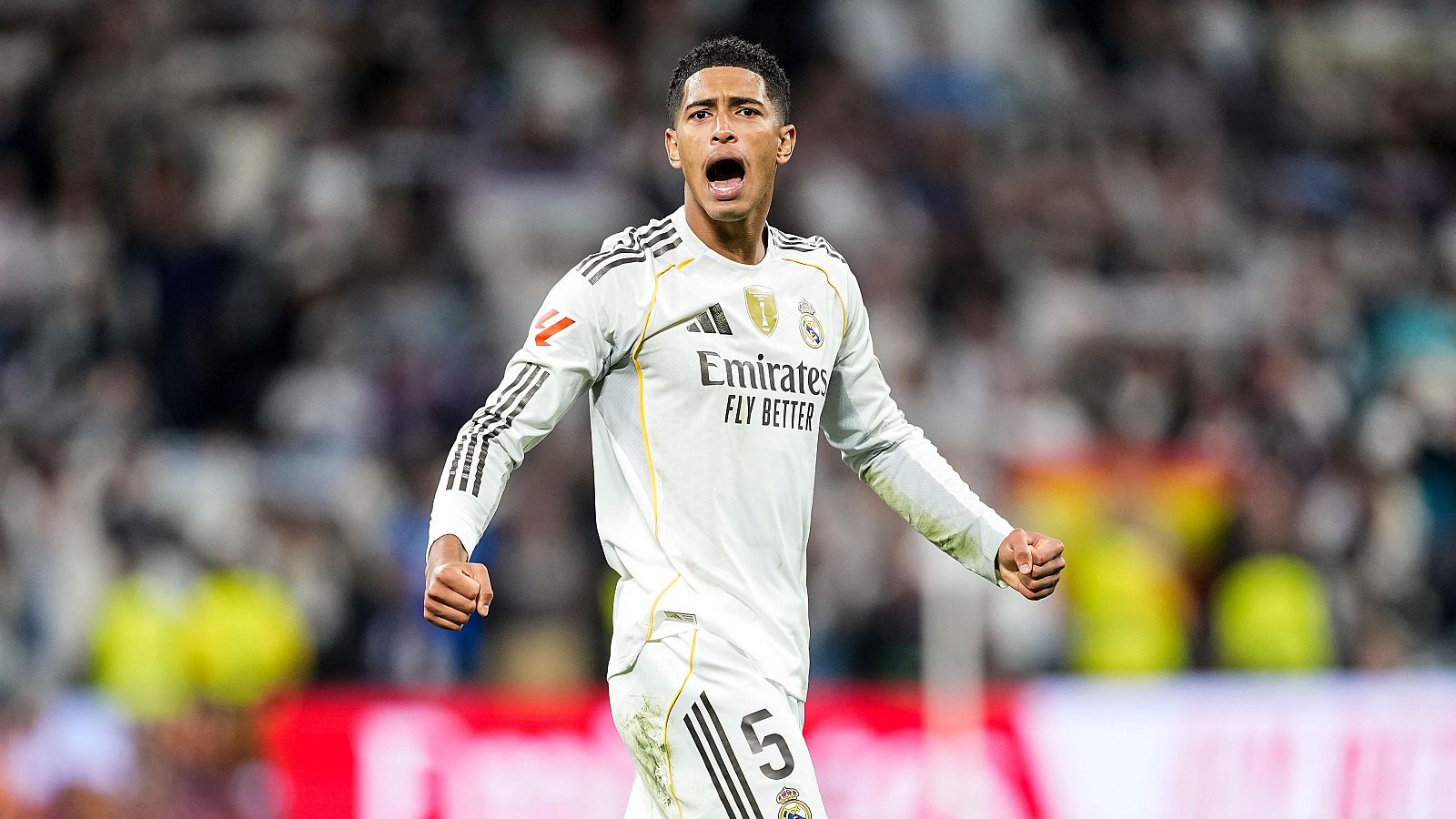Un jugador de fútbol, con la camiseta blanca de su equipo y el número 5, celebra un gol con los brazos en alto, ante un fondo difuminado con público.