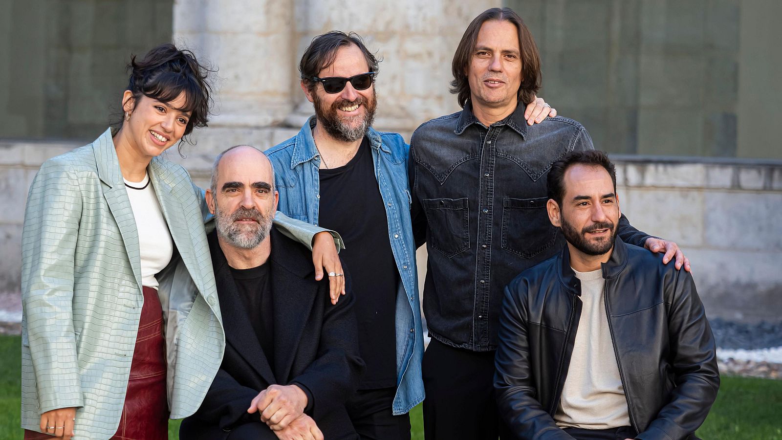 Un grupo de cinco personas posa frente a un edificio antiguo. Entre ellos, se observa a una mujer con chaqueta clara y falda roja, y varios hombres con diferentes estilos de vestimenta, incluyendo chaquetas de cuero y camisas vaqueras.