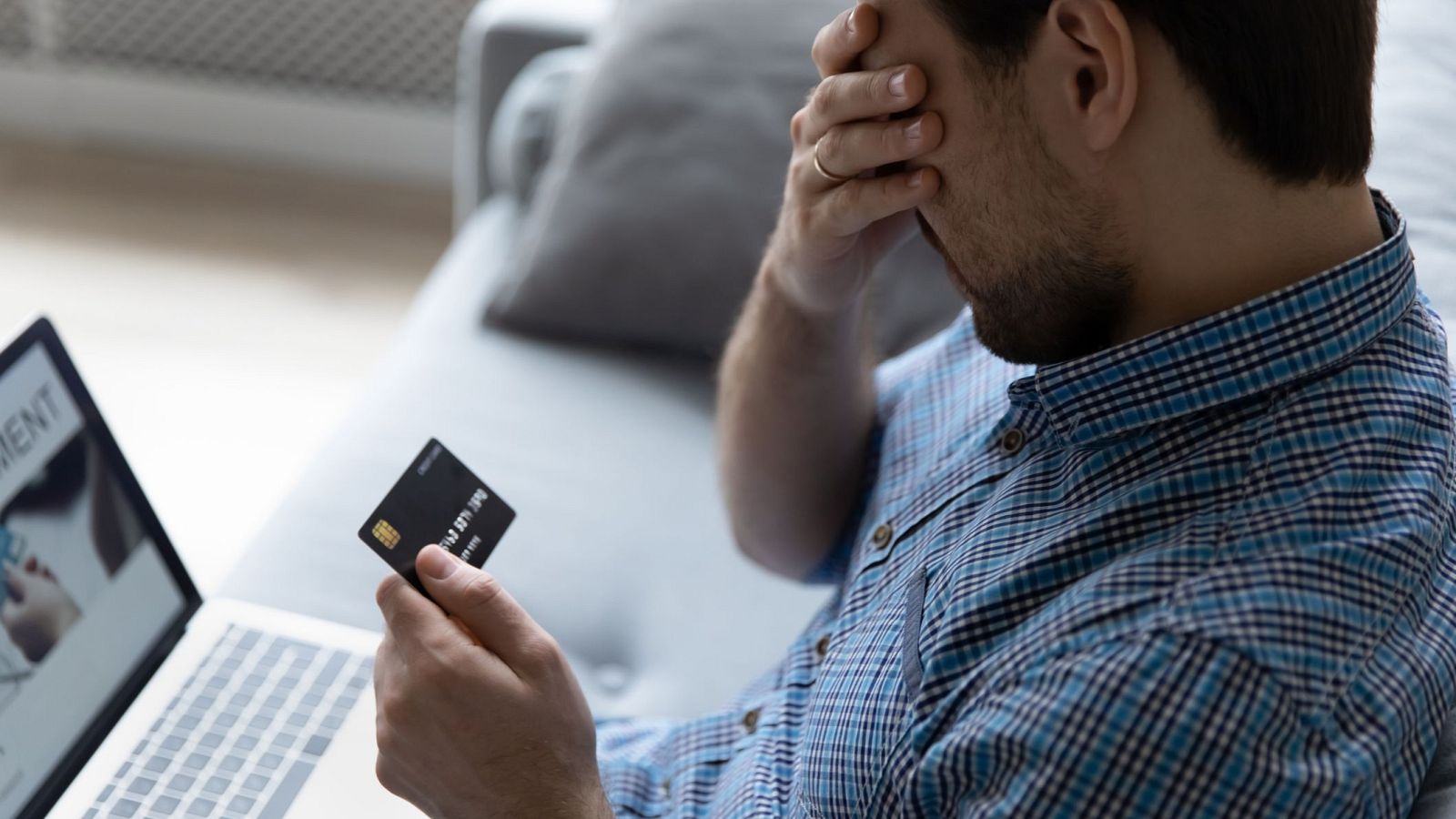 Un hombre barbudo, con camisa de cuadros, parece preocupado mientras está sentado en un sofá. Sostiene una tarjeta de crédito y frente a él hay un portátil abierto con la palabra "PAYMENT" en la pantalla.