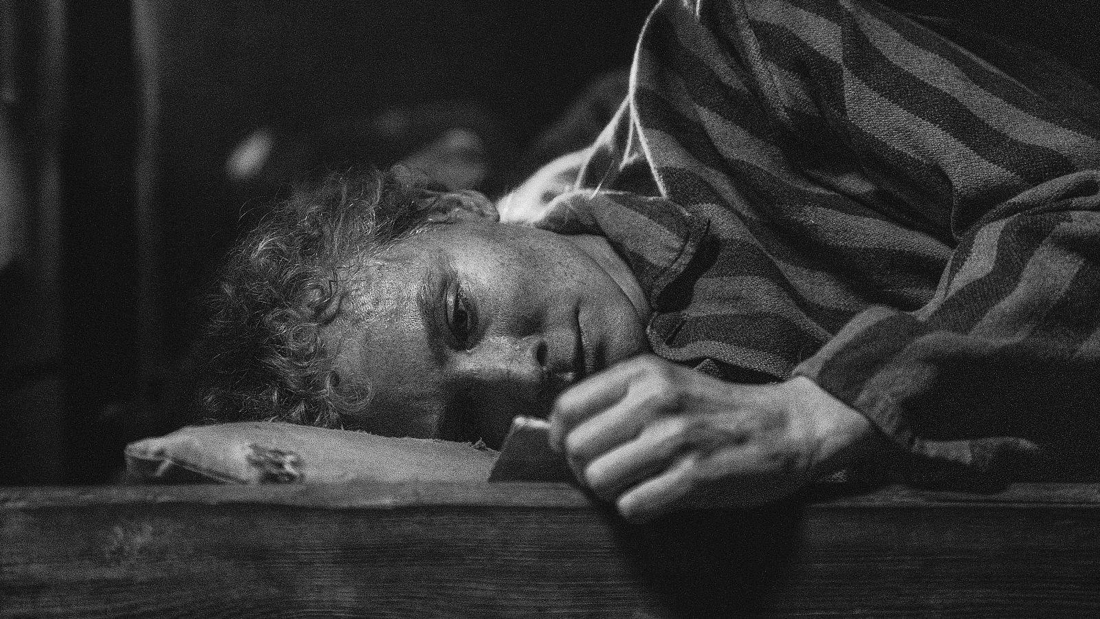 Diane Kruger, con una camisa a rayas, yace sobre una superficie de madera en una imagen en blanco y negro, mostrando una expresión de cansancio bajo una iluminación tenue.