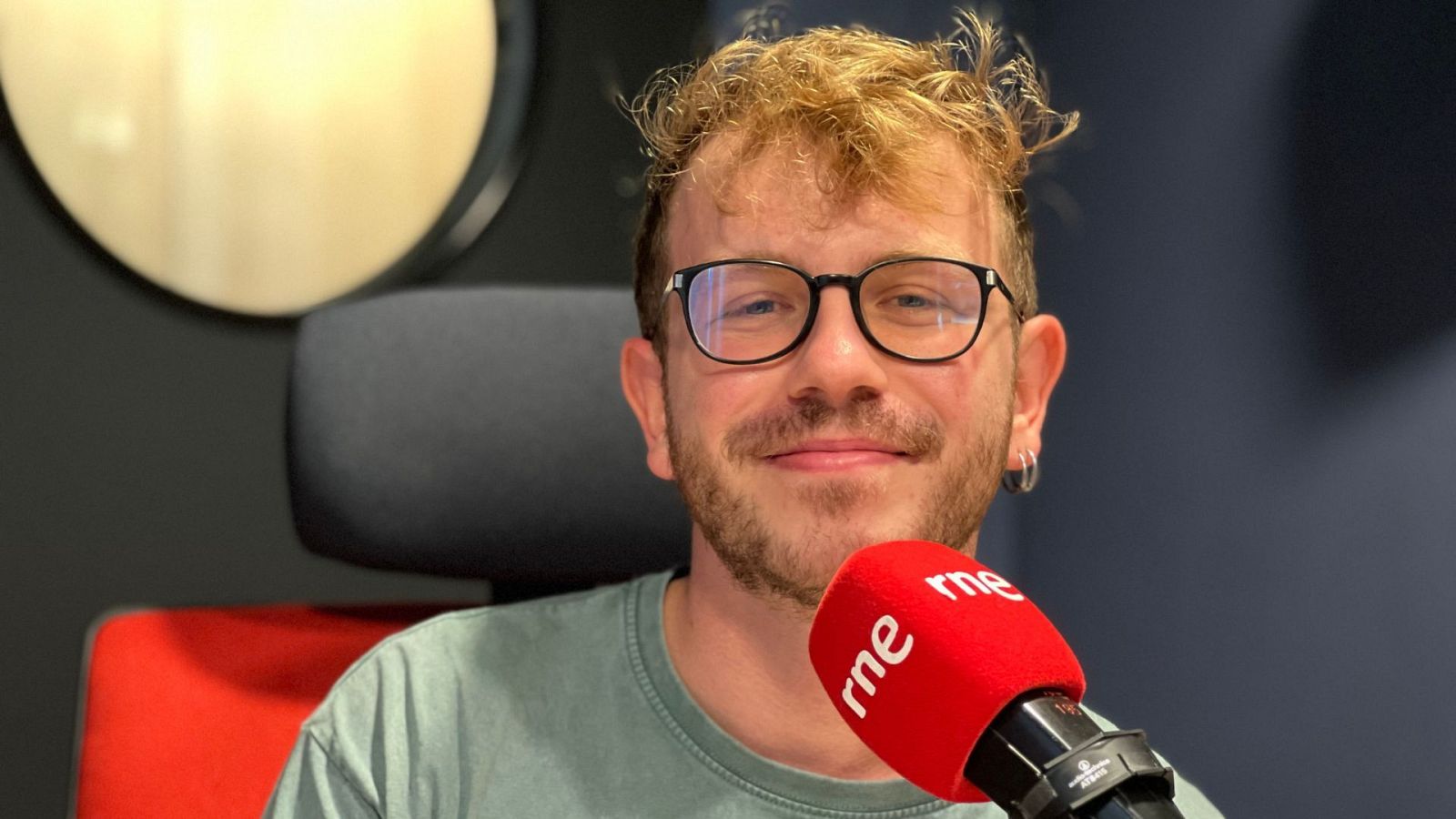 Nacho Roura, con gafas y camiseta verde, sonríe en un estudio de radio frente a un micrófono con el logo de una emisora. Se aprecia un pendiente en su oreja y un fondo oscuro.