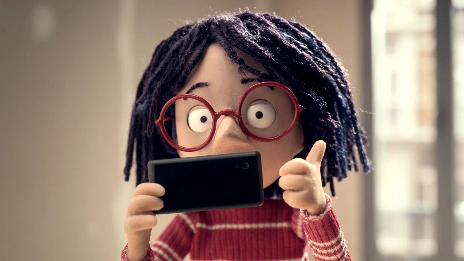 Una niña con gafas rojas y rastas, vestida con un jersey a rayas, sostiene un teléfono móvil negro con ambas manos, mostrando el pulgar hacia arriba.