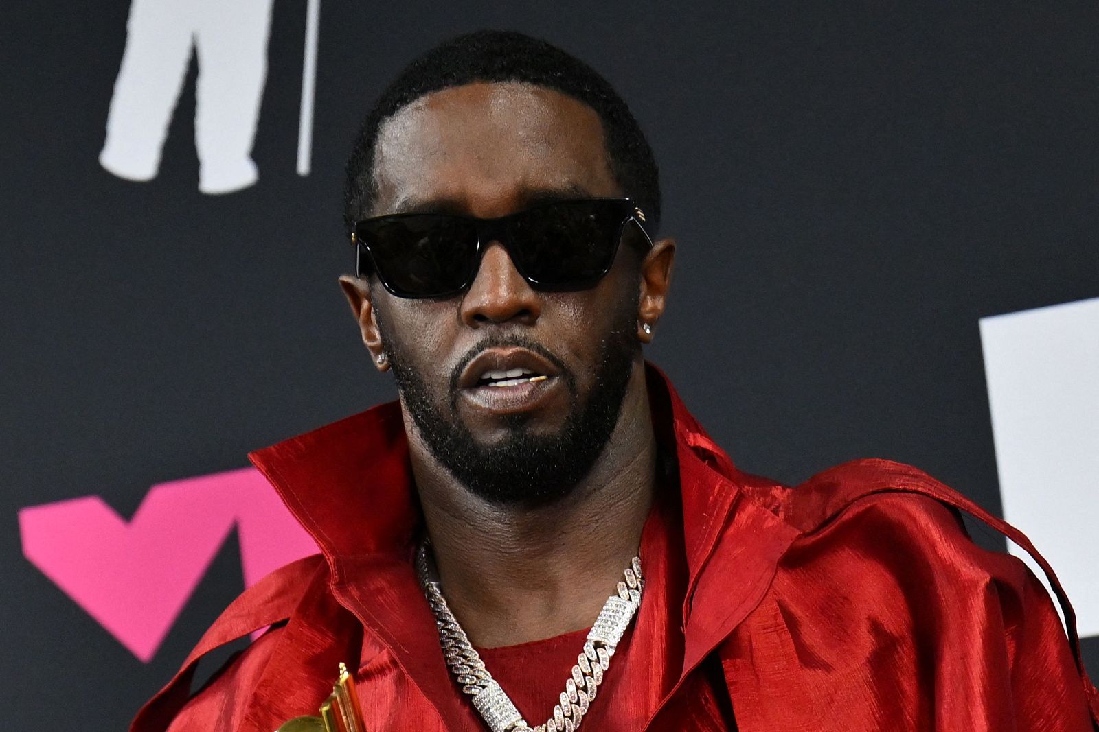 Diddy en los MTV Video Music Awards 2023, con camisa roja brillante, gafas de sol y collar de diamantes. Fondo negro con parte del logo de los premios.