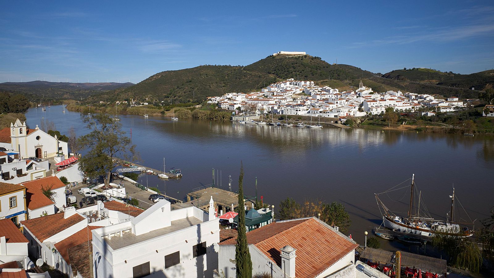 Una vista panorámica del río Guadiana en Portugal, con tejados anaranjados, una iglesia blanca y barcos en el río. Al otro lado, un pueblo de casas blancas se extiende por una colina, con una construcción en la cima.