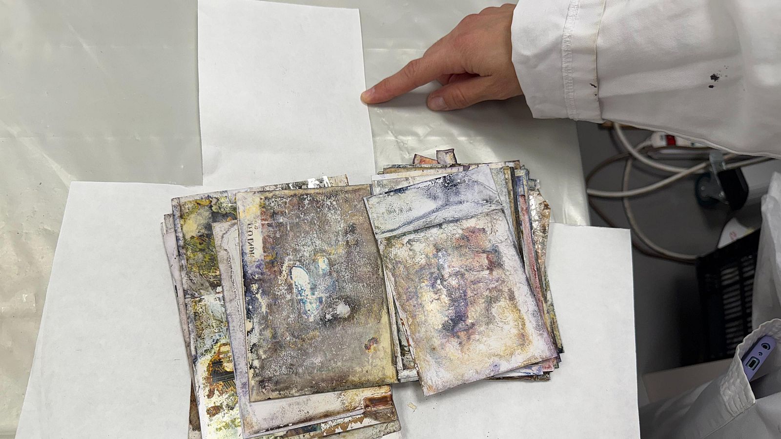 Un conjunto de fotografías dañadas, con manchas y suciedad, se apilan sobre una superficie clara. Una mano con bata blanca indica una hoja de papel en la escena.