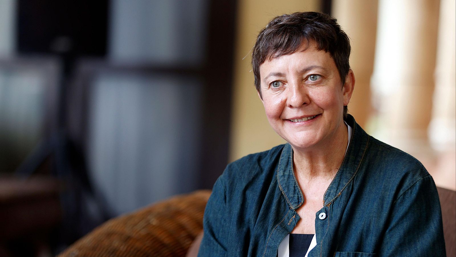 Eva Hache sonríe a la cámara, vestida con una camisa vaquera y una blusa. Se aprecia una columna y un objeto oscuro en el fondo, con una iluminación natural.