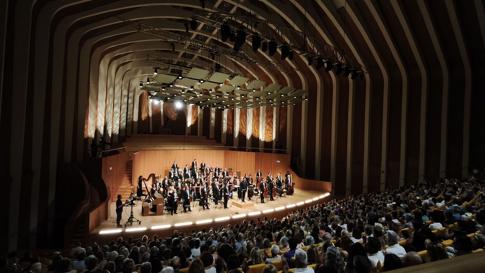 Una orquesta y coro, con vestimenta formal, se presenta en un auditorio moderno ante un público numeroso, interpretando música con instrumentos variados, como violines y arpas, en un evento en Valencia.