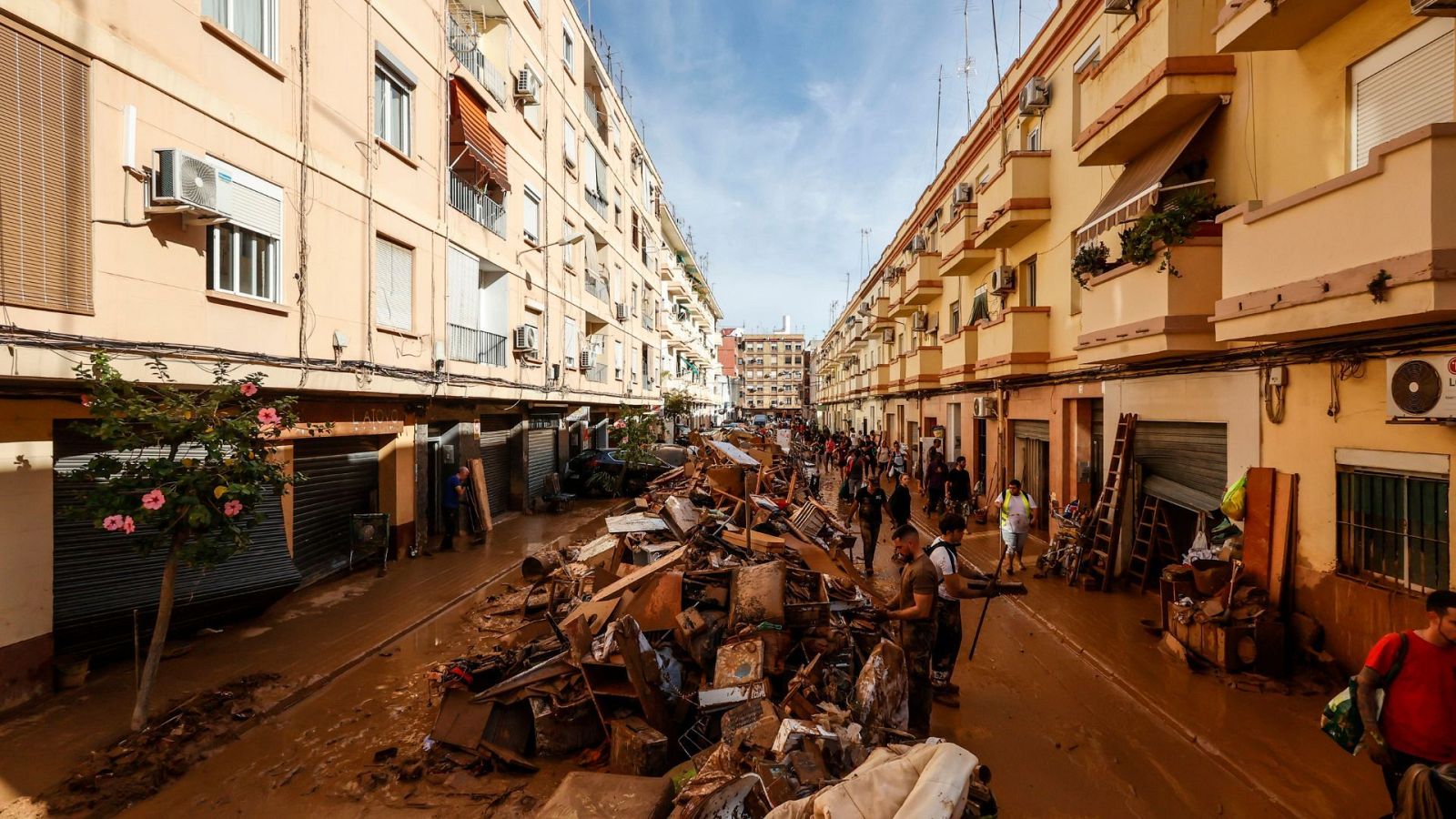 Una calle inundada con escombros y agua turbia muestra edificios y personas. Se observan locales con persianas bajas y un grupo de personas caminando y removiendo escombros en un día parcialmente nublado.