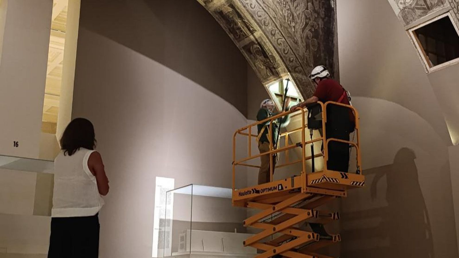 Técnicos en plataforma elevadora restaurando murales en un espacio con techo abovedado. Una persona observa el trabajo desde el suelo. Número "16" en la pared.