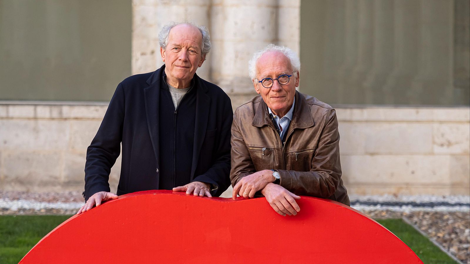 Dos hombres, uno con chaqueta oscura y otro con chaqueta de cuero marrón, posan frente a una estructura roja semicircular. Ambos tienen el pelo canoso y se encuentran en un entorno con una pared de piedra clara.