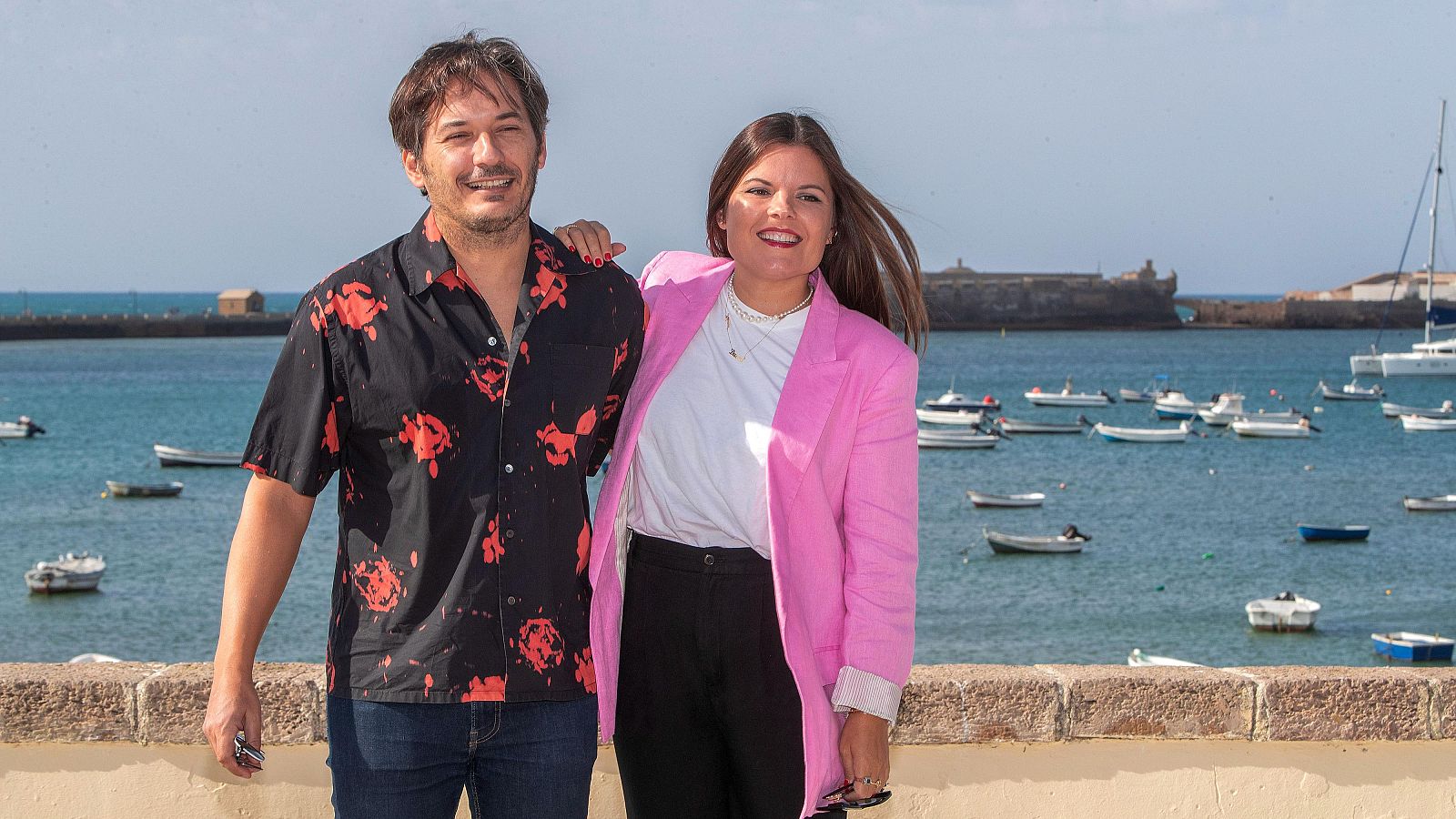 Dos hermanos posan frente a un paisaje marítimo. El hombre lleva una camisa estampada y vaqueros, mientras que la mujer viste una chaqueta rosa y pantalones oscuros, con su brazo sobre el hombro de su hermano.