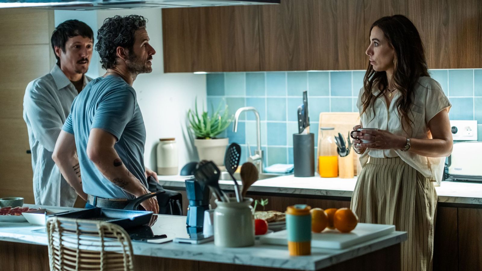 En una cocina con muebles de madera y azulejos azules, una mujer con una taza comparte espacio con dos hombres. Uno cocina con una sartén y camiseta azul, mientras el otro, con camisa blanca, observa.